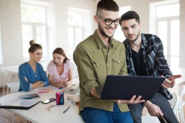 Gözlük ve gömlek sevinçle laptop ile modern ofis birlikte meslektaşları ile arka planda çalışan sakallı genç çocuklar. Grup yaratıcı insan iş yerinde vakit