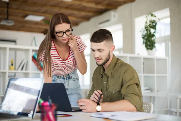 Young thoughtful man in shirt and woman in striped T-shirt and eyeglasses dreamily working together with laptop. Creative business people spending time at work in modern cozy office