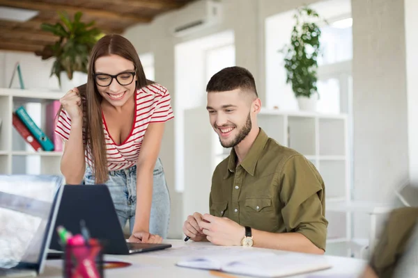 Young joyful man in shirt and woman in striped T-shirt and eyeglasses happily working together with laptop. Creative smiling business people spending time at work in modern office