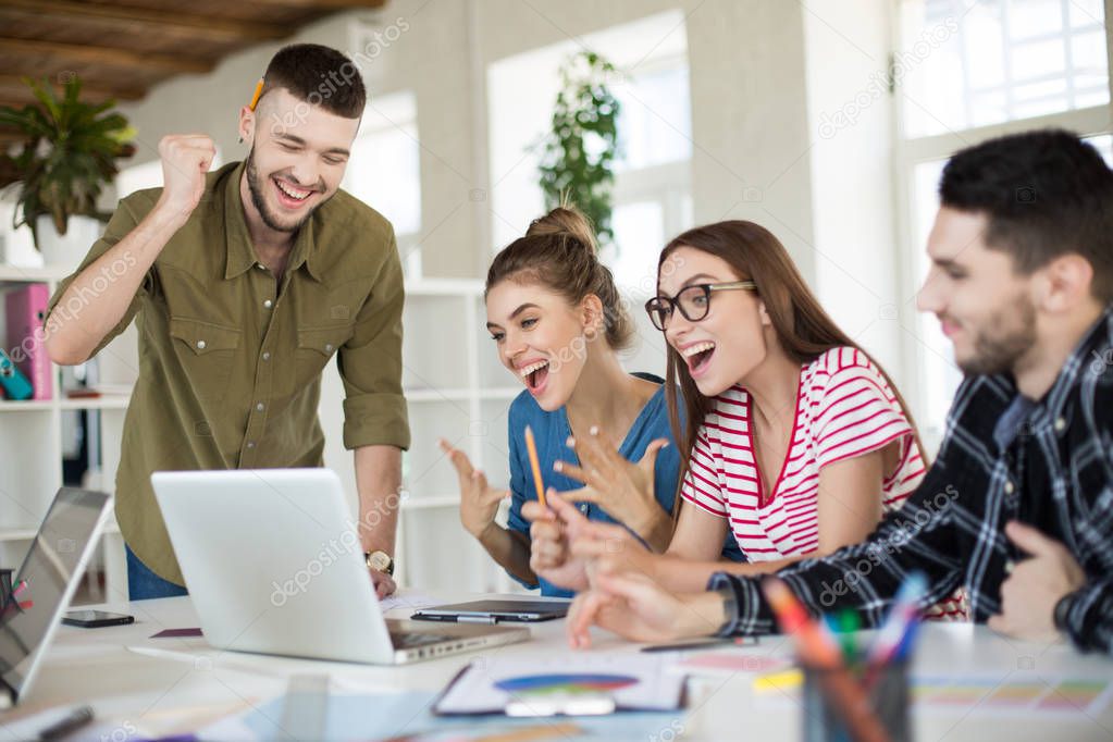 Young emotional business people joyfully working on laptop together. Group of laughing men and women spending time at work in modern cozy office