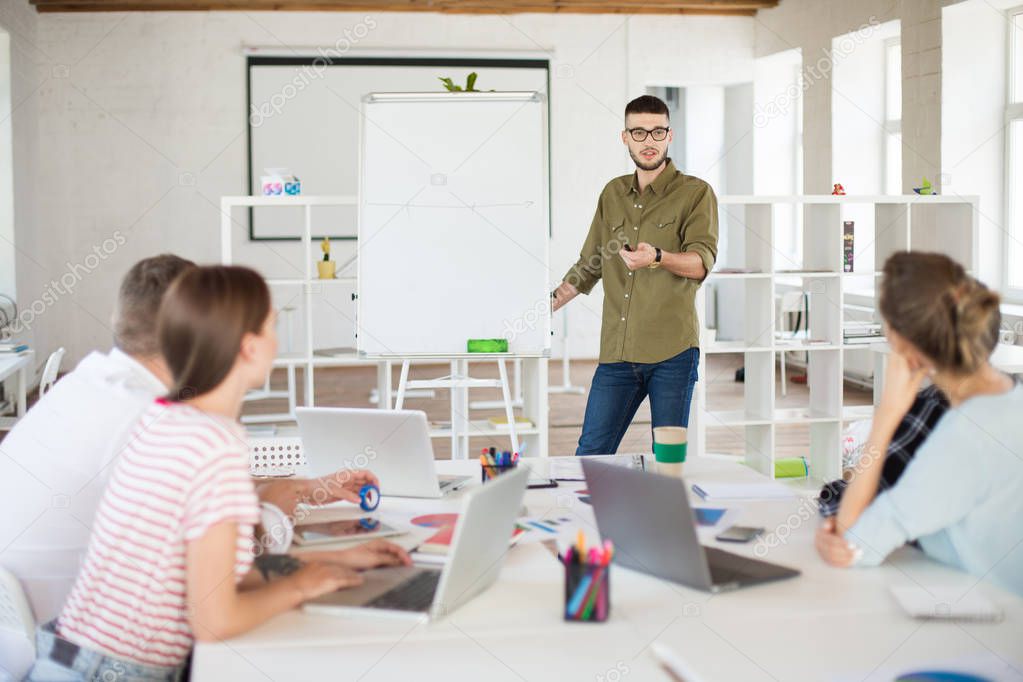 Young man in eyeglasses and shirt standing near board discussing new project with colleagues at work. Group of people working together in modern office