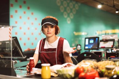 Young upset female cashier in uniform thoughtfully looking in camera working in modern supermarket clipart