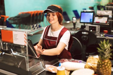 Pretty smiling female cashier in uniform happily using cashbox working in modern supermarket clipart