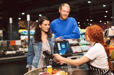 Mutlu bir şekilde modern süpermarkette kredi kartıyla ürünler için ödeme kasiyer Resepsiyon yakınındaki güzel gülümseyen çift ayakta