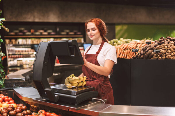 Young smiling seller in apron behind counter with vegetables weighing bananas on scale in modern supermarket
