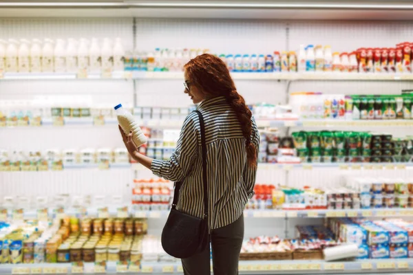 漂亮的女孩在眼镜和条纹衬衫选择牛奶在现代超市乳制品部门 — 图库照片