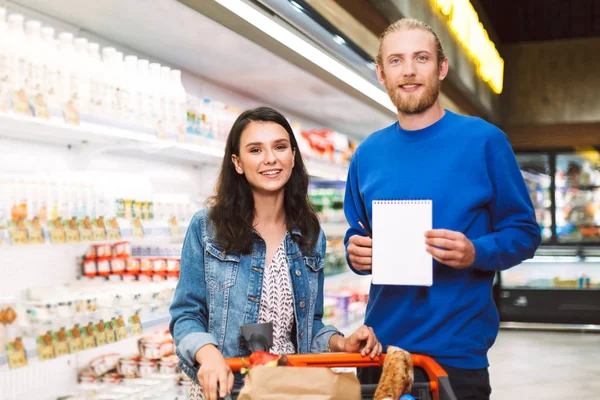 年轻的微笑夫妇与购物单和手推车快乐地看着相机在现代超市乳品部 — 图库照片