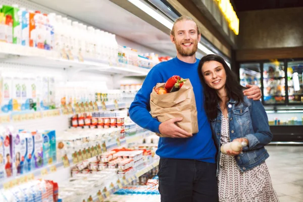 美丽的笑脸夫妇与纸杂货袋充满产品快乐地看着相机在现代超市乳制品部门 — 图库照片