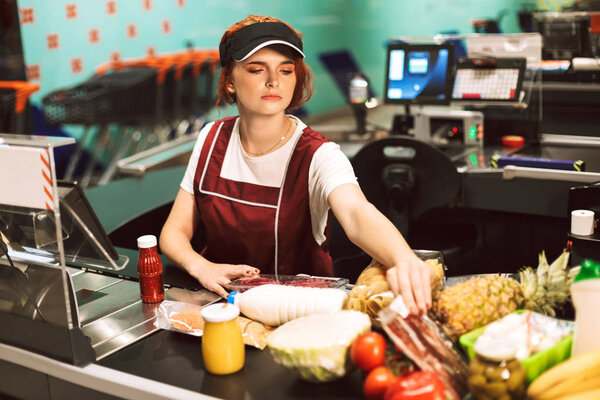 Young female cashier in uniform thoughtfully looking on products working in modern supermarket
