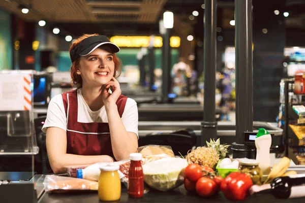 年轻开朗的女收银员高兴地看着一旁的产品在现金线附近的现代超市工作 — 图库照片