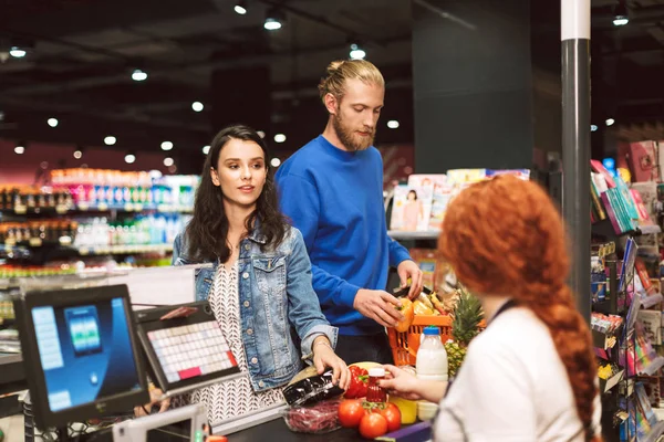 年轻美丽的夫妇站在收银柜台附近购买产品在现代超市 — 图库照片