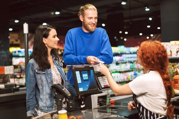 年轻欢快的夫妇站在收银台附近愉快地给出纳员在现代超市的信用卡 — 图库照片