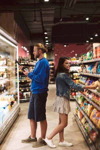 Young guy and pretty girl standing back to back choosing products spending time in modern supermarket