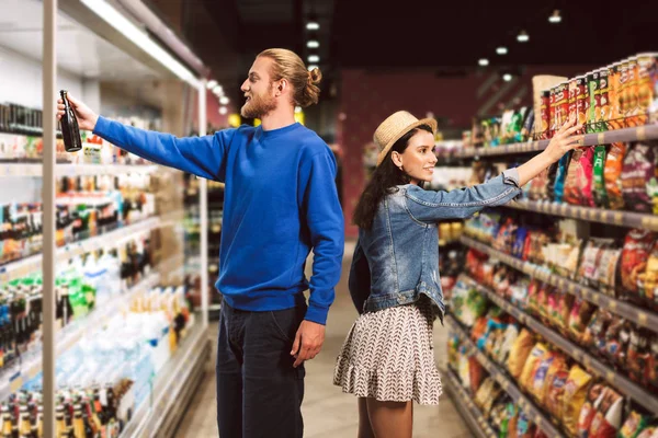 年轻的微笑的家伙选择啤酒 而漂亮的女孩接近选择芯片在现代超市 — 图库照片
