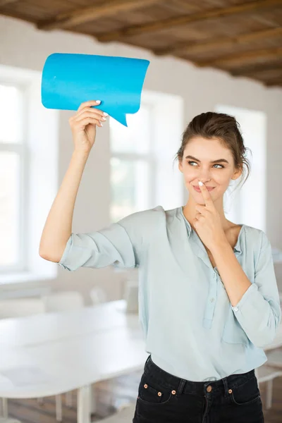 Young beautiful woman in blouse dreamily looking aside holding blue paper shape of message in hand at work in modern empty office