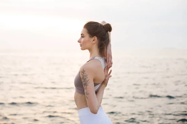 Beautiful girl in sporty top and white leggings holding hands behind back with sea view on background. Young woman dreamily looking aside practicing  yoga by the sea