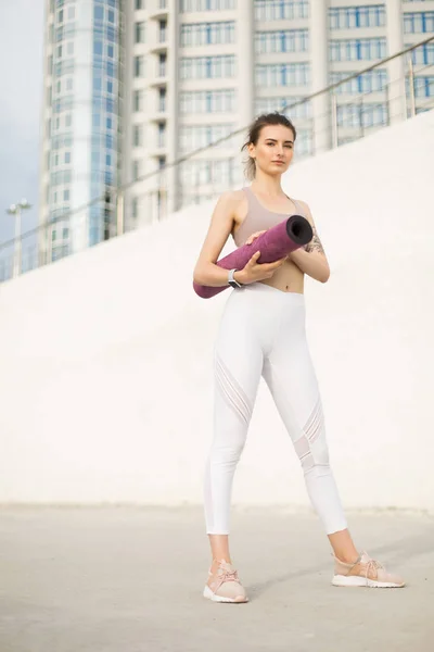 Young beautiful plus size woman in sporty top and leggings with red  headphones sitting on knees on yoga mat while thoughtfully looking aside in  city park Stock Photo