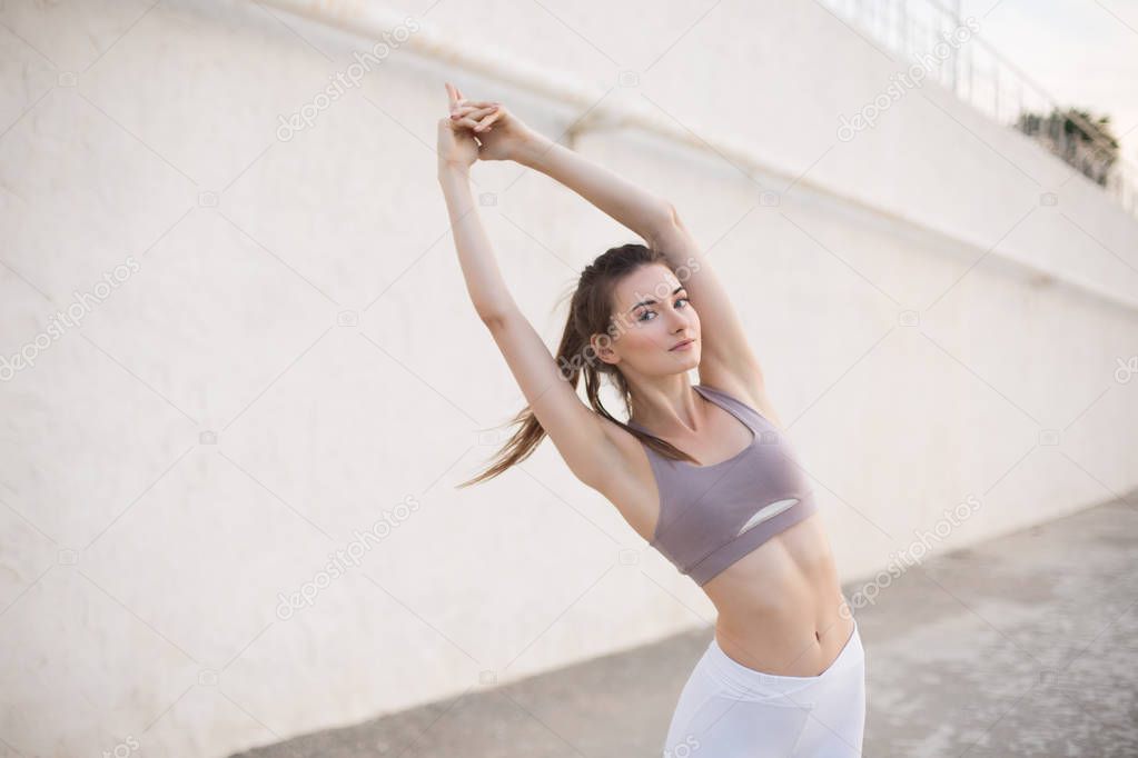 Beautiful girl in sporty top stretching thoughtfully looking in camera. Young woman training alone outdoor