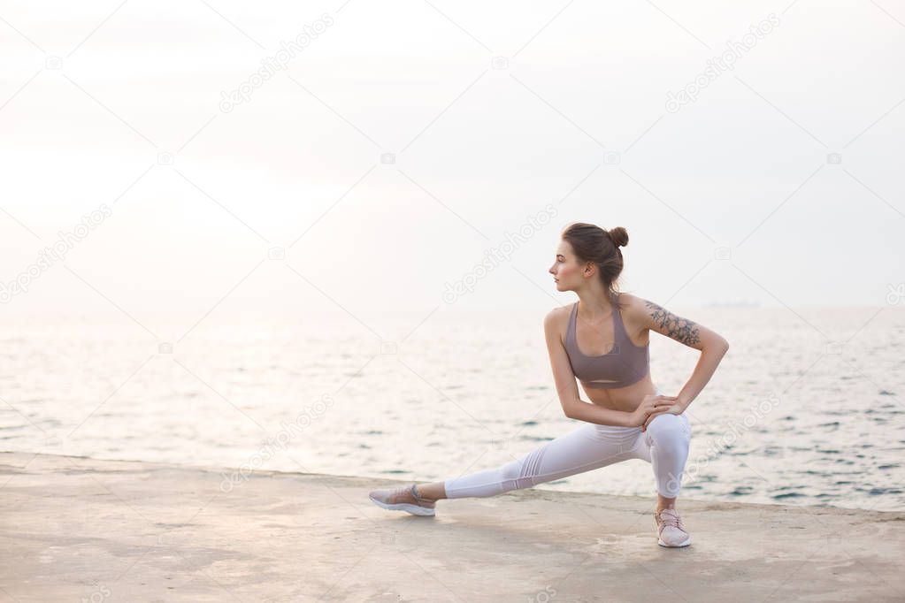 Thoughtful girl in sporty top and white leggings practicing yoga with amazing sea view on background. Young woman stretching by the sea