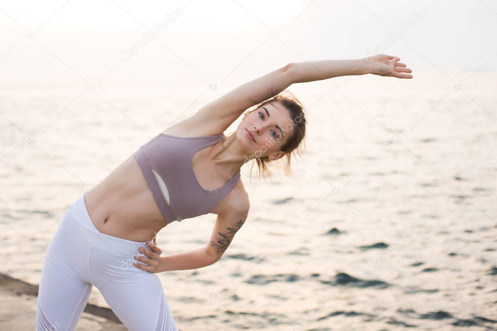 Pretty girl in sporty top and white leggings dreamily looking in camera practicing yoga with sea view on background. Young woman stretching by the sea