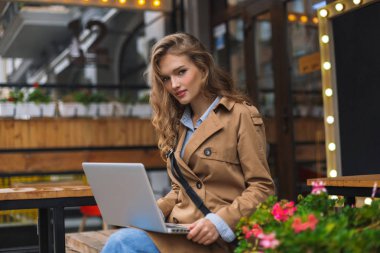 Trençkot sersem kamera laptop ile oturma seyir genç çekici kız vakit açık kafe Teras diz.