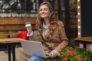 Trençkot mutlu kamera seyir holding kredi kartı el ile laptop diz açık kafe terasında oturan genç gülümseyen kadın