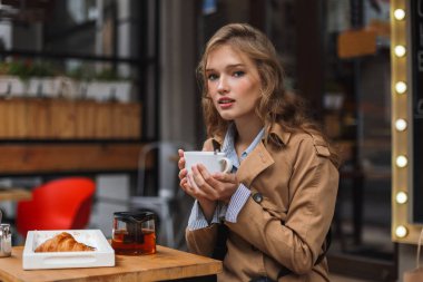 Trençkot sersem kamera tutarak Çaydanlık ve beyaz Kupası rahat kafe Teras, açık arıyor genç çekici kadın