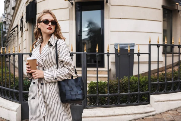 美丽的女孩在条纹的风衣和太阳镜与黑色手提包拿着咖啡去在手若有所思地看一边走在舒适的城市街附近 — 图库照片