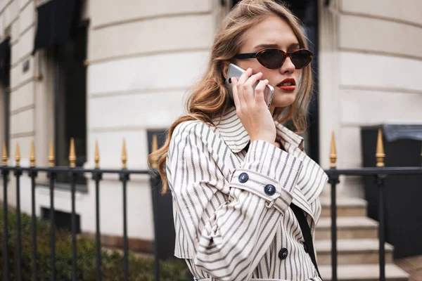 Joven Mujer Seria Gabardina Rayada Hablando Pensativamente Teléfono Celular Caminando — Foto de Stock