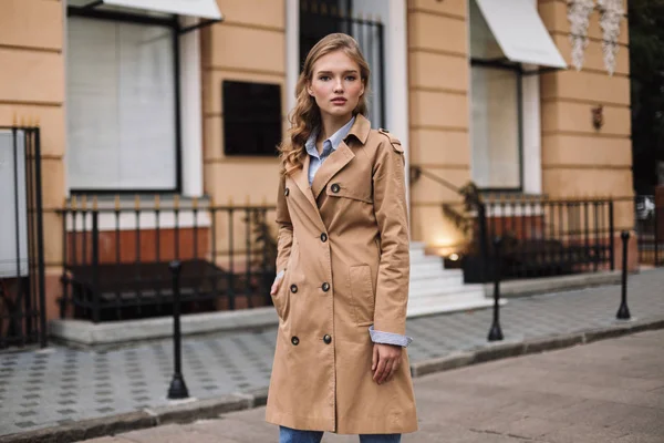 Young attractive woman in trench coat thoughtfully looking in camera walking around cozy city street