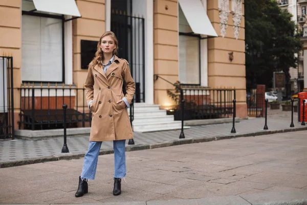 美丽的沉思的女孩在战壕大衣和牛仔裤若有所思地看着相机漫步在舒适的城市街道 — 图库照片