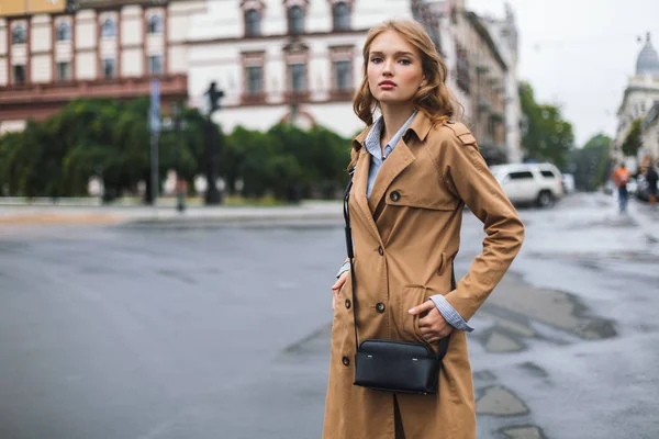 Young attractive woman in trench coat with little black cross bag dreamily looking in camera spending time on cozy city street