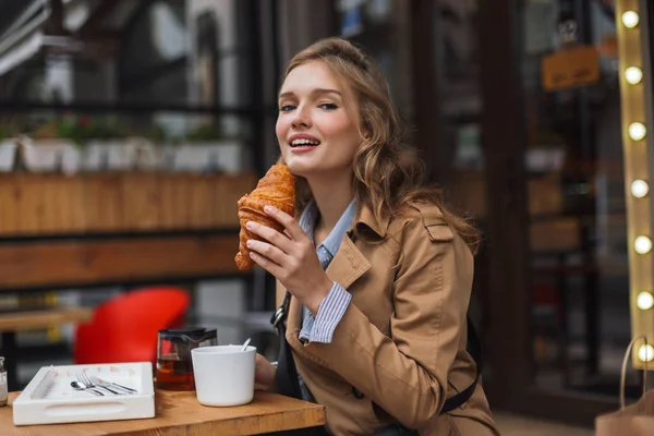 Young beautiful woman in trench coat dreamily looking in camera eating croissant spending time outdoor at cozy cafe terrace