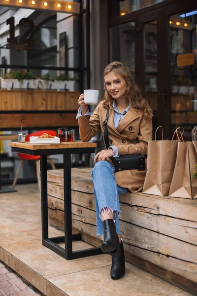 トレンチ コートやお茶の居心地の良いカフェのテラスで過ごす時間屋外が手に白い保持カップ探してカメラでうっとりとジーンズで美しい少女 — ストック写真