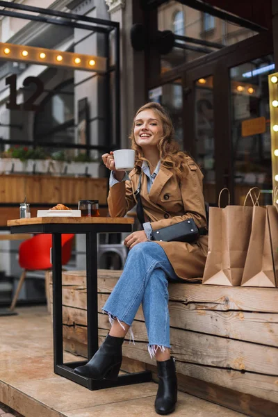 トレンチ コートとジーンズ持株白一杯のお茶の居心地の良いカフェのテラスで過ごす時間屋外が手に探してカメラで嬉しそうで笑顔美人 — ストック写真