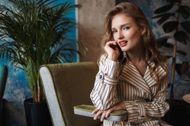 Çizgili trençkot sersem seyir fotoğraf makinesindeki harcama zaman modern kafede dalgalı saçlı genç gülümseyen güzel kadın