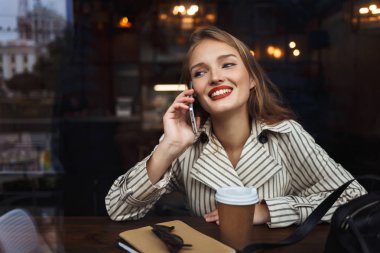 Oldukça modern rahat kafe zaman harcama yakınındaki gitmek çizgili trençkot sevinçle bir kenara seyir fincan kahve ile cep telefonu konuşurken kız gülümseyerek