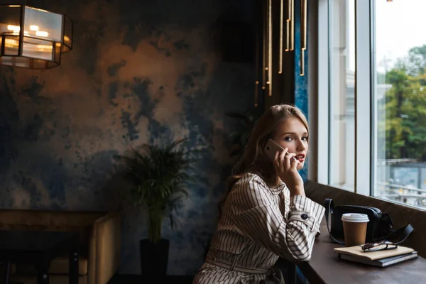 Çizgili trençkot düşünceli seyir fotoğraf makinesindeki harcama zaman modern kafede büyük pencere ile cep telefonu konuşurken genç güzel kadın