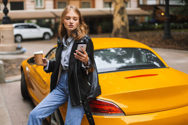 Young attractive woman in leather jacket leaning on yellow sport car holding cup of coffee to go in hand thoughtfully using cellphone on city street