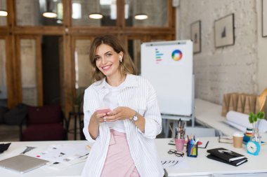 Beyaz gömlek ve radyo earphones sevinçle modern rahat ofis içinde işte cep telefonu kullanarak kamera seyir masada yaslanmış güzel gülümseyen kadın