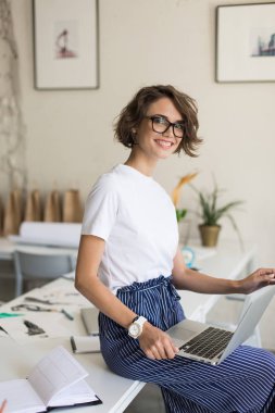 Gözlük ve mutlu bir şekilde iş yerinizde modern ofis dizüstü bilgisayar üzerinde çalışan kamera seyir masasında oturan beyaz tişört kısa kıvırcık saçlı oldukça neşeli kız