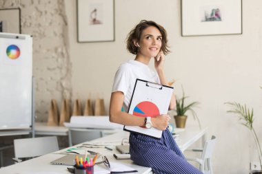 Radyo earphones sersem bir kenara holding klasör diyagramı ile modern rahat Ofis çalışmalarında bakarak masasında oturan kısa kıvırcık saçlı genç gülümseyen kadın