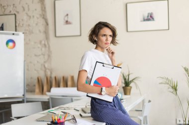 Kablosuz kulaklık ne yazık ki bir kenara holding klasör diyagramı ile modern ofis çalışmalarında bakarak masasında oturan kısa kıvırcık saçlı Genç ciddi kadın