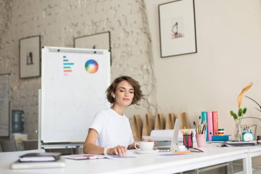 Radyo earphones ve beyaz tişört sersem iş yerinizde modern rahat office kahve fincan ile Laptop çalışma kısa kıvırcık saçlı güzel kız