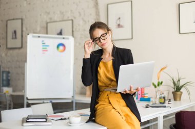 Gözlük, siyah ceket ve radyo earphones üstünde laptop modern ofis içinde işte kahve ile çalışma masasında oturan genç düşünceli kadın
