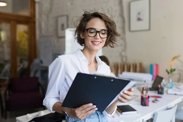 年轻美丽的微笑的女孩 短卷发在眼镜和白色衬衫手持文件夹在现代舒适的办公室工作的手 — 图库照片