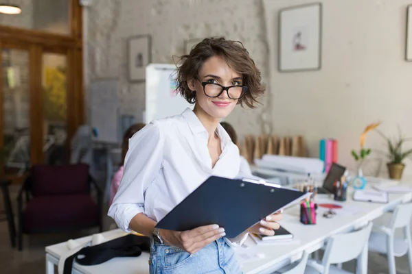 年轻漂亮的女孩与短卷曲的头发在眼镜和白色衬衫持有文件夹在手朦胧地看着相机在现代舒适的办公室工作 — 图库照片