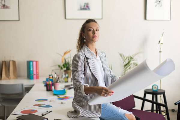 Молодая серьезная женщина в полосатой куртке и джинсах сидит на столе задумчиво глядя в камеру, держа в руках большую бумагу как человек в руках на работе в современном офисе
