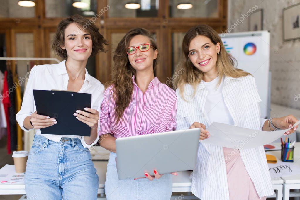 Group of young beautiful stylish women with laptop and folder happily looking in camera working together in modern cozy office