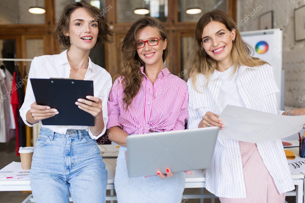 Group of young beautiful stylish women with laptop and folder joyfully looking in camera working together in modern cozy office. Pretty girls spending time at work